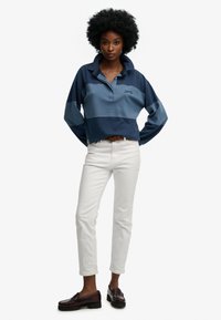 Kobieta z afro w fryzurze, ubrana w granatowo-niebieską koszulkę polo z długim rękawem, białe spodnie, brązowy pasek i ciemnobrązowe mokasyny, stojąca z rękami na biodrach.