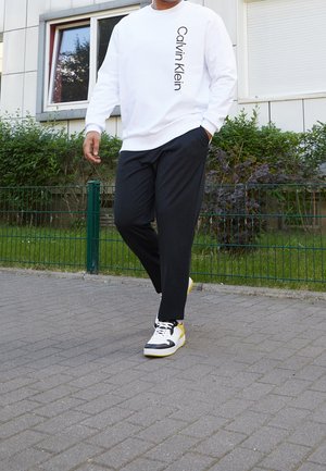 Person, der går på fortovet iført hvid Calvin Klein sweatshirt, sorte bukser og hvide sneakers med gule og sorte detaljer.