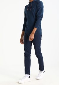 Felpa blu navy con tasca frontale e coulisse, abbinata a pantaloni slim-fit blu navy e sneakers bianche. Texture liscia, design casual.