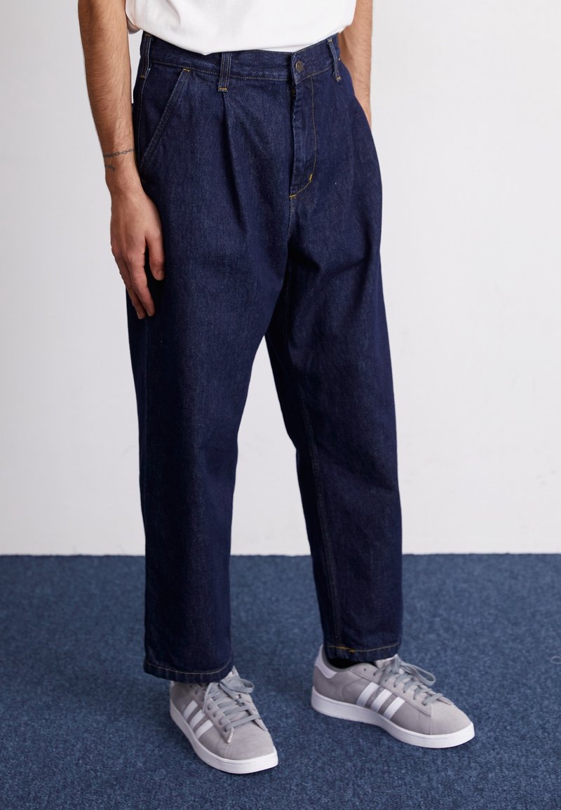 Ciemnoniebieskie dżinsy o luźnym kroju, z zakładkami, zapięciem na guziki i widocznymi przeszyciami. Połączone z szarymi trampkami.