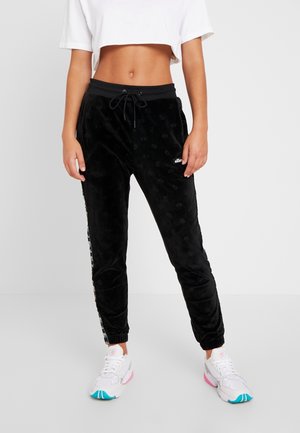 Kobieta ubrana w czarne welurowe spodnie dresowe z trokami w pasie i logo, biały krótki t-shirt oraz białe sneakersy z różowymi i turkusowymi akcentami.