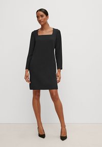 Schwarzes Langarmkleid mit quadratischem Ausschnitt, taillierter Silhouette und glatter Textur, das bis zur Mitte des Oberschenkels reicht, kombiniert mit spitzen schwarzen Absatzschuhen.