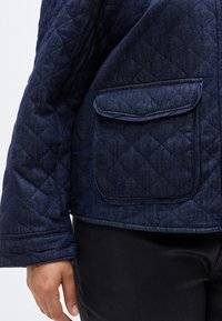 Πουκάμισο από τζιν με καπιτονέ ύφασμα σε σκούρο μπλε, με τσέπη στο στήθος, φαρδιές μανσέτες και ομαλή υφή. Το κάτω άκρο είναι ίσιο.