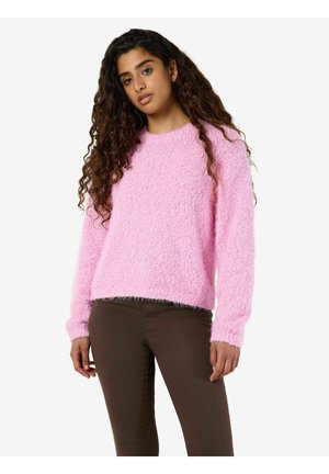 Femme aux longs cheveux bouclés portant un pull rose pelucheux et un pantalon marron foncé, debout devant un fond blanc.