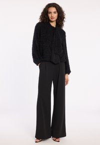 Schwarzer, flauschiger Pullover mit einer Schleife am Hals, kombiniert mit weitgeschnittenen schwarzen Hosen. Das Outfit hat eine weiche Textur und eine lässige Silhouette.