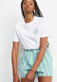Hvit bomulls T-skjorte med en sirkulær lyseblå logo på brystet, kombinert med grønne og blå stripete shorts med elastisk liv. 