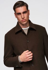 Ruskea villakangastakki, jossa on jämäkkä kaulus, yksinappinen suljenta ja istuva leikkaus, puettuna vaaleanharmaan neuleen päälle. Sileä pinta, minimalistinen muotoilu.