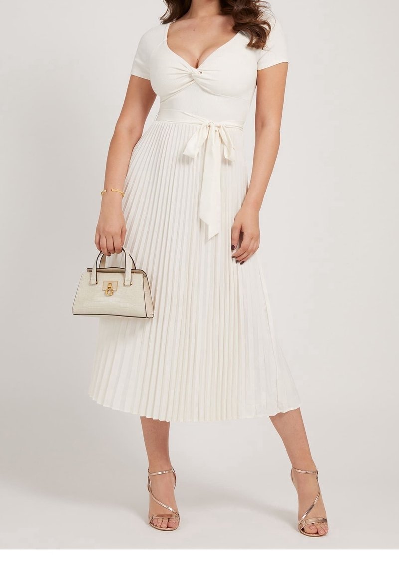 Femme en robe midi blanche plissée avec une ceinture à la taille, tenant un petit sac à main beige, portant des sandales dorées à talons hauts avec des brides.