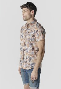 Hombre con camisa de manga corta con estampado tropical abotonada y pantalones cortos de mezclilla arremangados, de pie de lado frente a un fondo liso.