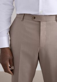 Pantalons ajustés beiges avec une texture lisse, devant plat et fermeture à un bouton. Associés à une chemise blanche à manches longues.