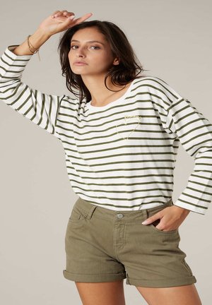 Femme posant avec le bras levé, portant un haut à manches longues à rayures blanches et vertes et un short olive retroussé, sur un fond neutre.