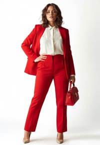 Roter maßgeschneiderter Anzug mit langer Hose und Blazer, kombiniert mit weißem Hemd und goldfarbenen Absatzschuhen. Hält eine kleine rote Handtasche.