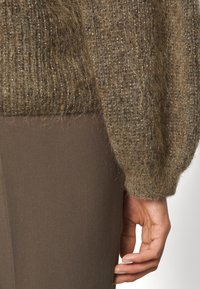 Gros plan d'une main à côté d'une manche de pull en maille brune texturée et d'un pantalon brun lisse.