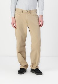 Beige Cargohosen mit geradem Schnitt, ausgestattet mit Seitentaschen und einem Knopfverschluss, gefertigt aus Baumwolle oder einem ähnlichen Stoff. Schwarze Sneakers getragen.