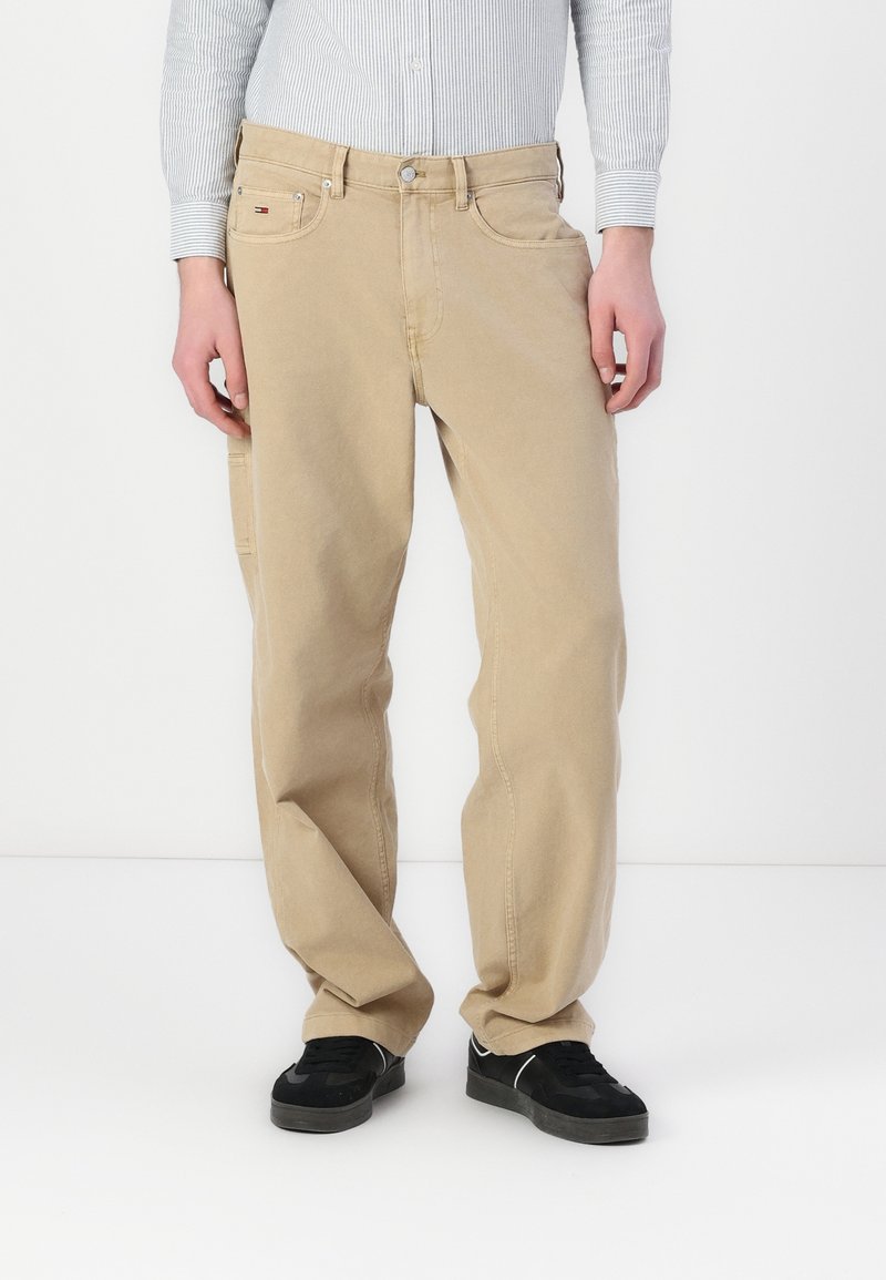 Beige Cargohosen mit geradem Schnitt, ausgestattet mit Seitentaschen und einem Knopfverschluss, gefertigt aus Baumwolle oder einem ähnlichen Stoff. Schwarze Sneakers getragen.