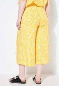 Gelbe Culotte-Hosen mit einem weißen abstrakten Muster, ausgestattet mit einem elastischen Bund. Kombiniert mit schwarzen Rutsch-Sandalen mit einer strukturierten Sohle.