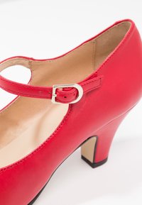 Chaussure Mary Jane en cuir rouge avec un bout arrondi, une bride avec une boucle argentée et un talon haut effilé. Texture lisse et design minimaliste.