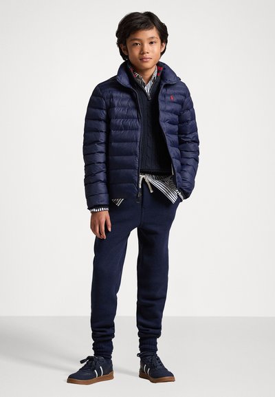 Marineblå pufferjakke med sydd mønster, lagd over en marineblå strikkegenser, stripete skjorte, marineblå joggebukser og marineblå joggesko med hvite detaljer.