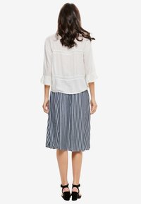 Blouse blanche bordée de dentelle avec des manches mi-longues jusqu'au coude, associée à une jupe à rayures bleu marine et blanches longueur genou, portée avec des sandales noires à fines lanières.