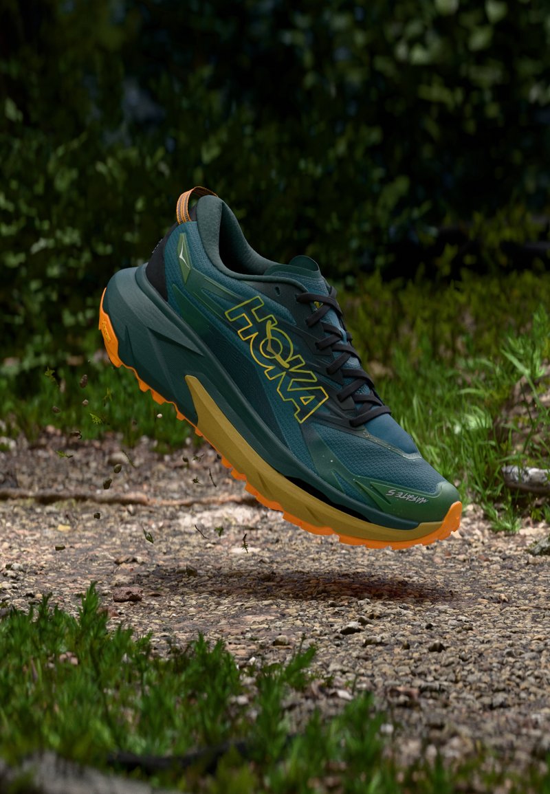 Πράσινο αθλητικό παπούτσι με μαύρες λεπτομέρειες και πορτοκαλί εξωτερική σόλα. Διαθέτει αναπνεύσιμο επάνω μέρος από δίχτυ, υποστηρικτικό σχεδιασμό και τον εμφανή λογότυπο "HOKA".