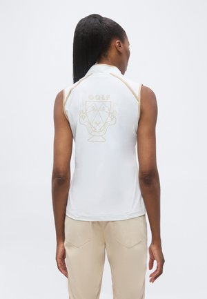 Tumehobusega naine kannab valget varrukateta golfi särki beežide äärte ja embleemiga, kombineeritud beežide pükstega, seistes seljaga vaataja poole.