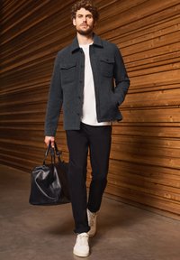 Grå texturerad jacka med två framfickor, vit t-shirt, svarta byxor, vita sneakers och en svart läderväska mot en träbakgrund.