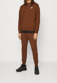 Osoba stojící oblečená v hnědém svetru Nike a odpovídajících teplákách s černými teniskami na jednobarevném pozadí.