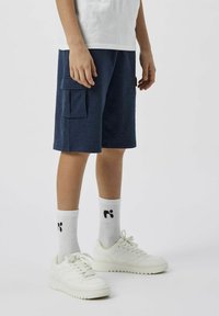 Giovane che indossa pantaloni cargo blu scuro, calze bianche a manica corta con logo nero, scarpe da ginnastica bianche e una maglietta bianca, in piedi su uno sfondo neutro.