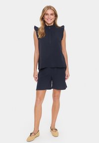 Tumši zila divdaļīga tērpa komplekts, kas sastāv no bezpiedurkņu tops ar volānu pleciem, pogu aizdari un atbilstošām biksēm no strukturētas auduma.