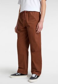 Pantalons en coton marron avec taille élastique et cordon de serrage, offrant une coupe décontractée et des poches latérales. Associés à des baskets noires et blanches.