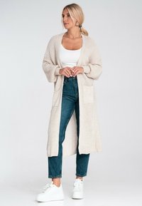 Beżowy sweter dzianinowy z długimi rękawami, otwarty z przodu, z dwiema przednimi kieszeniami, noszony na białym topie, w połączeniu z niebieskimi dżinsami i białymi trampkami.