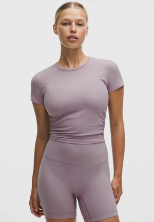 Mujer con camiseta ajustada de manga corta color lavanda y pantalones cortos ciclistas de talle alto, de pie frente a un fondo liso gris claro.