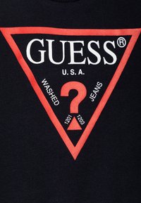 Svart t-skjorte med en rød trekantet logo. Logoen inkluderer teksten "GUESS U.S.A." og "WASHED JEANS", sammen med tall og et spørsmålstegn-design.