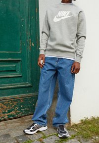 Sudadera gris con gran logo blanco de Nike, jeans sueltos azules y zapatillas deportivas negras y plateadas, de pie frente a una puerta verde.