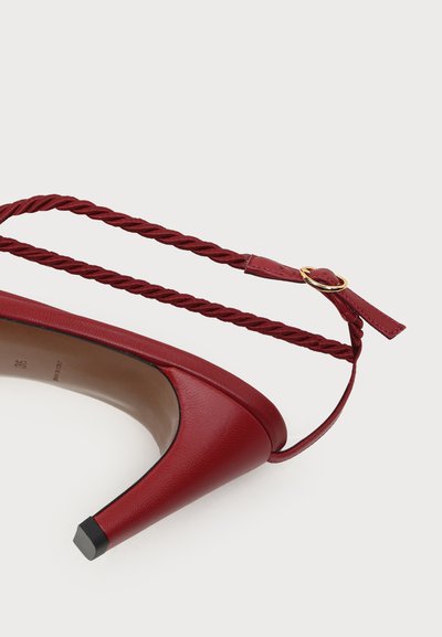 Chaussure à talon haut rouge avec bride de cheville torsadée et boucle dorée, présentée sur fond blanc uni.