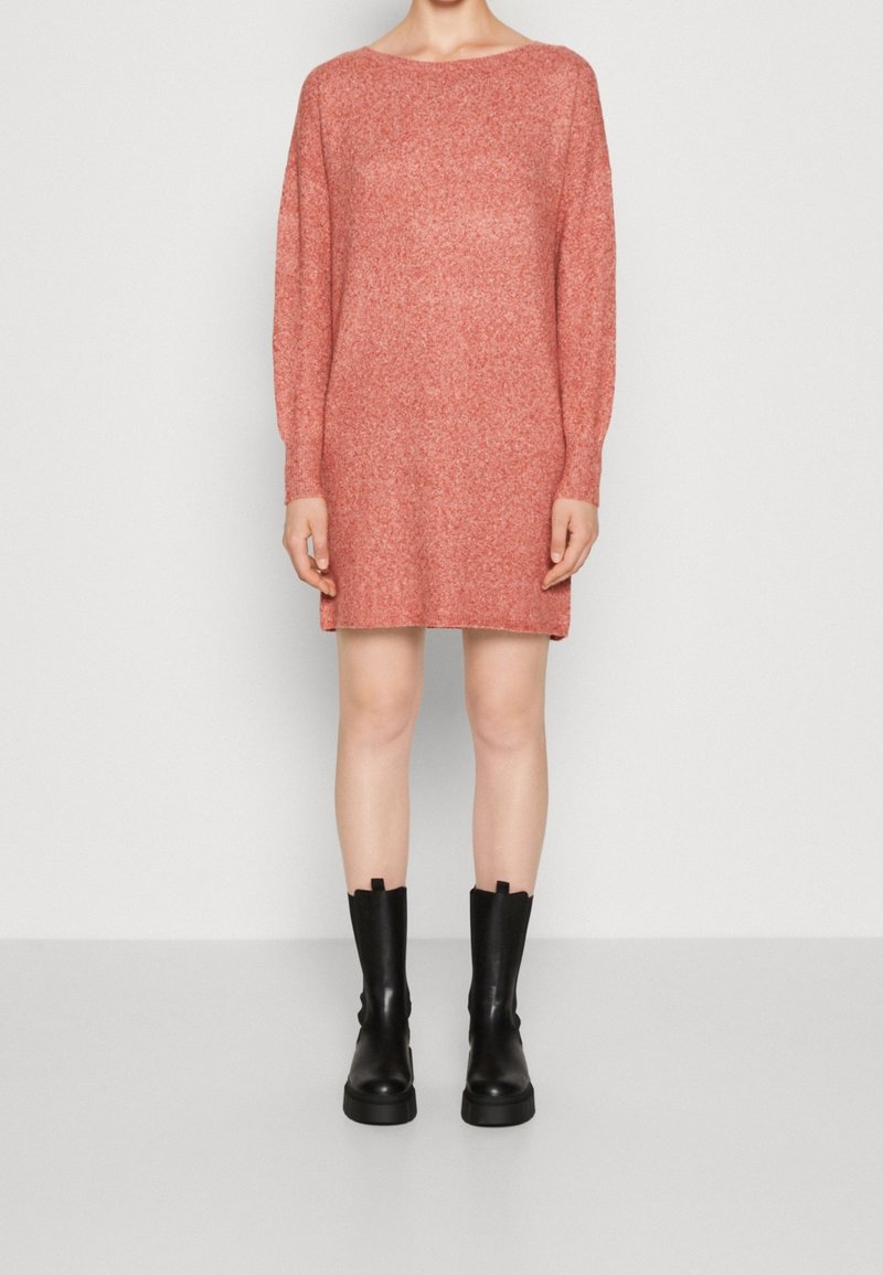 Personne portant une robe en maille rose à manches longues, longueur genou, et des bottes noires épaisses mi-mollet, debout sur un fond uni.