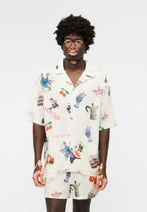 Homme atteint de vitiligo portant une chemise blanche à manches courtes et un short assorti avec des motifs colorés sur le thème des vacances, debout devant un fond uni.