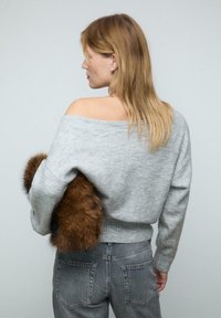 Pull en laine gris au design off-shoulder, avec un ourlet côtelé et des manches longues, accompagné d'un jean gris foncé et d'un accessoire en fausse fourrure marron.