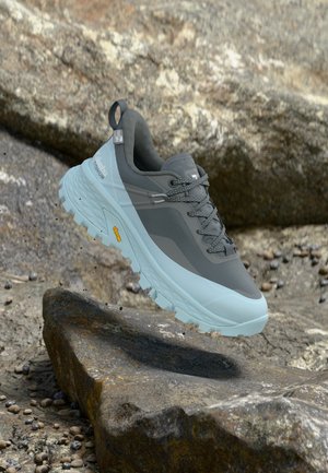 Γκρι και ανοιχτό μπλε πεζοπορικό παπούτσι Columbia Titanium, αιωρούμενο πάνω από βραχώδες έδαφος, που παρουσιάζει ανθεκτική σόλα Vibram και ανθεκτικό σχεδιασμό.