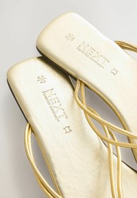 Sandali piatti dorati con texture in pelle liscia, caratterizzati da sottili cinturini incrociati e logo del marchio impresso nella soletta. Taglia 6.5/40.
