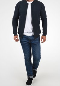 Tmavě námořnická bunda na zip s žebrovanou texturou, doplněná bílým tričkem a modrými džínami. Outfit je dokončen černými teniskami.