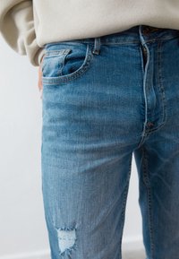 Vaqueros de mezclilla azul claro con corte ajustado, que presentan un parche desgastado en el muslo derecho y un diseño clásico de cinco bolsillos.