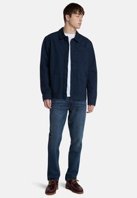 Chaqueta de algodón azul marino con cierre de botones en el frente, dos bolsillos laterales y un bolsillo en el pecho, combinada con jeans azules y zapatos marrones de cordones.