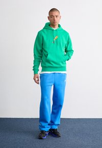 Hanorac verde cu buzunar frontal și detalii grafice colorate, purtat cu pantaloni de trening blue deschis și adidași negri.