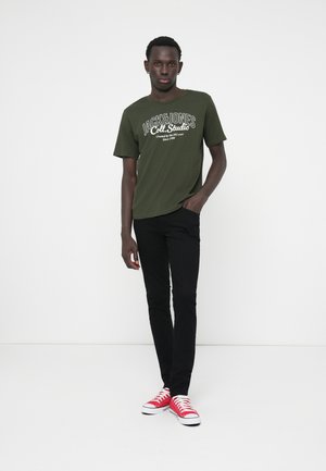Mørkegrønn T-skjorte med hvitt logo og tekst, sorte slim-fit bukser, røde sneakers med hvite såler, som står på en ensfarget bakgrunn.