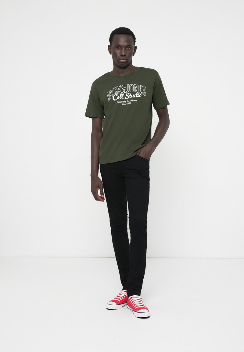 Dark green T-shirt with white logo and text, black slim-fit pants, red sneakers with white soles, standing on a plain background.