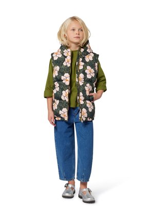 Mouwloze vest met bloemenpatroon in het groen met roze bloemen, gecombineerd met een groene blouse met lange mouwen en blauwe spijkerbroeken. Zilveren schoenen maken de look compleet.