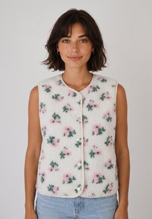 Donna con capelli corti e scuri indossa un gilet smanicato bianco in pile con motivi floreali rosa e verdi e jeans azzurri su sfondo uniforme.