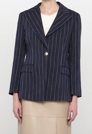 Blazer bleu marine à fines rayures avec bouton argenté, larges revers, poches à rabat, porté sur un top en maille claire et une jupe beige.