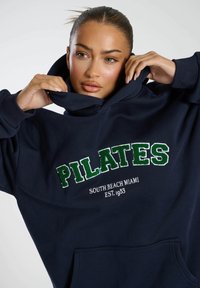 Felpa blu navy in morbido tessuto con la scritta "PILATES" in ricamo verde, e "SOUTH BEACH MIAMI EST. 1953" in bianco sotto.